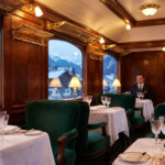 Épopée historique du wagon-restaurant Orient Express Intérieur luxueux d'un wagon-restaurant Orient Express avec des sièges en velours vert et une vue sur des montagnes enneigées.