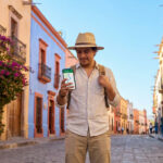 Sécurité au Mexique en 2026 : conseils aux voyageurs Un voyageur avec un chapeau et un sac à dos consulte son itinéraire sur son téléphone dans une rue colorée au Mexique.