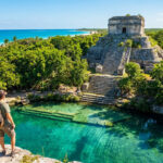 Un voyageur avec sac à dos contemple un ancien temple maya situé au bord d'un cénote turquoise et de la mer des Caraïbes.
